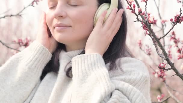 En ung kvinna lyssnar på musik på hörlurar och mediterar, hon är i en blommande trädgård på våren. Ett underbart ögonblick av avkoppling och lugn. Fred och enhet med naturen. — Stockvideo