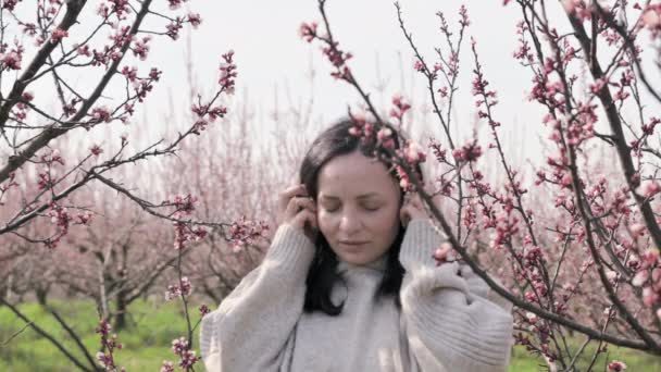 Mladá žena poslouchá hudbu na sluchátkách a medituje, na jaře je v kvetoucí zahradě. Nádherný okamžik odpočinku a klidu. Mír a jednota s přírodou. — Stock video