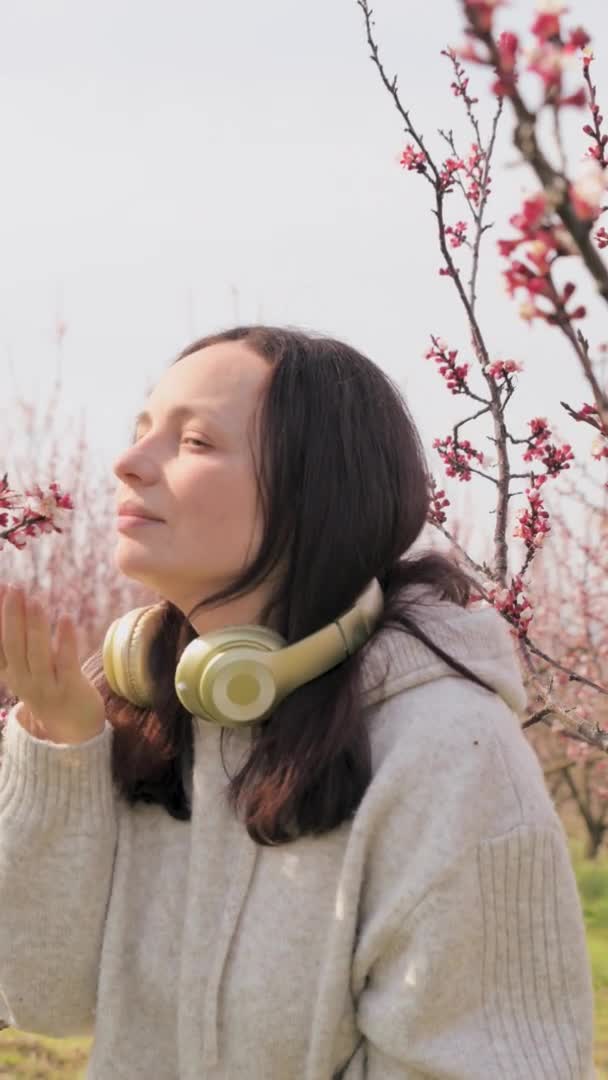 Een jonge vrouw luistert naar muziek op de koptelefoon en mediteert, ze is in het voorjaar in een bloeiende tuin. Een heerlijk moment van ontspanning en rust. Vrede en eenheid met de natuur. — Stockvideo