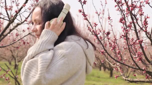 Une jeune femme écoute de la musique sur écouteurs et médite, elle est dans un jardin fleuri au printemps. Un merveilleux moment de détente et de tranquillité. Paix et unité avec la nature. — Video