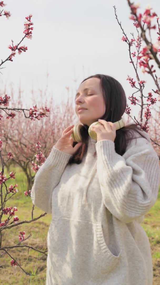 Uma jovem ouve música em fones de ouvido e medita, ela está em um jardim florescente na primavera. Um momento maravilhoso de relaxamento e tranquilidade. Paz e unidade com a natureza. — Vídeo de Stock