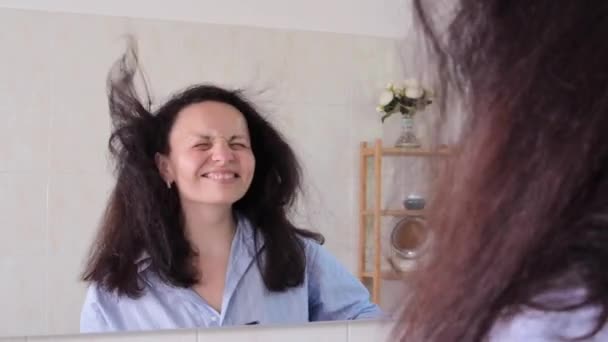 Une femme joyeuse sèche ses cheveux, elle s'amuse et se regarde dans le miroir. Prendre soin de son apparence à la maison — Video