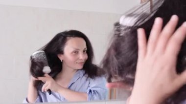 Bir kadın saçını saç kurutma makinesiyle kurutur. Uzun saçlı esmer bir kadın banyoda saçlarıyla ilgileniyor. Bir insanın aynadaki yansıması. Yaşam tarzı videosu. Yüksek kalite 4k görüntü