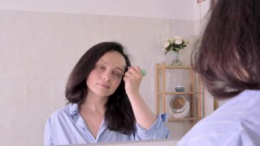 Banyoda makyaj yapan bir kadın. Avrupalı görünümlü genç esmer bir kadın banyoda onun yüzüne bakar. Aynaya yansıyor ve makyaj fırçası kullanıyor. 4k görüntü