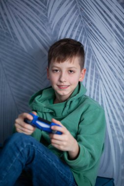 Kanepedeki çocuk bilgisayar oyunu oynuyor. Çocuk bir oturma odasında elinde joystick tutarak gülümsüyor..