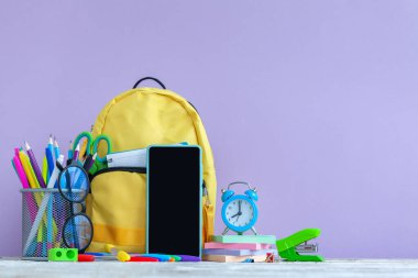 Okula dönüş kavramı. Tam sarı okul çantası, akıllı telefon, gözlük, çalar saat ve mor arka planda kırtasiye malzemesi..