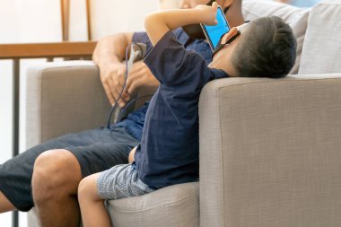 Asyalı bir çocuğun yüz maskesi takıp koltukta akıllı telefon oynaması. Cep telefonu olan küçük çocuk kafede koruyucu maske takıyor. Hastalık salgını sırasında halka açık yerleri ziyaret etme kavramı. Yeni normal.