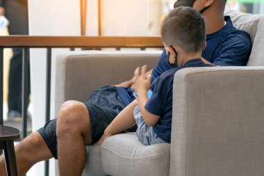 Asyalı bir çocuğun yüz maskesi takıp koltukta akıllı telefon oynaması. Cep telefonu olan küçük çocuk kafede koruyucu maske takıyor. Hastalık salgını sırasında halka açık yerleri ziyaret etme kavramı. Yeni normal.