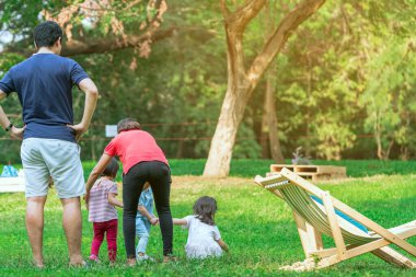 Asyalı anne ve babanın arka manzarası yeşil bahçedeki çimenlikte kızlarına iyi bak. Güzel bir aile anı. Mutlu aile, yeşil doğada birlikte vakit geçiriyor. Anneye odaklan..