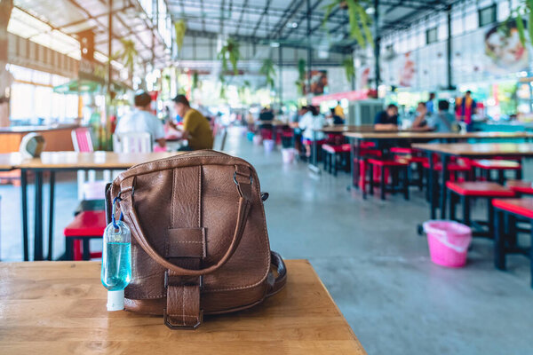 Mini portable blue alcohol gel bottle to kill Corona Virus(Covid-19) hang on brown leather shoulder bag of a woman on table at cafeteria. New normal lifestyle. Selective focus on alcohol gel.