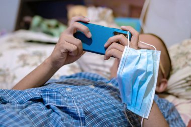 Coronavirus (C0VID-19) enfeksiyonunu önlemek için cerrahi bir maske, cep telefonlarıyla oyun oynamak için genç bir adamın elinde asılı duruyordu. Yeni normal yaşam tarzı. Cerrahi maskeye odaklan.