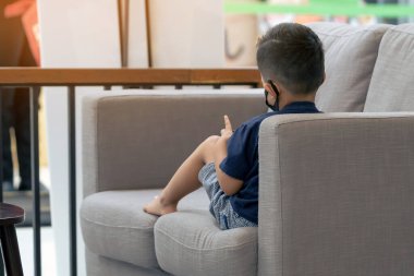 Asyalı bir çocuğun yüz maskesi takıp koltukta akıllı telefon oynaması. Cep telefonu olan küçük çocuk kafede koruyucu maske takıyor. Hastalık salgını sırasında halka açık yerleri ziyaret etme kavramı. Yeni normal.