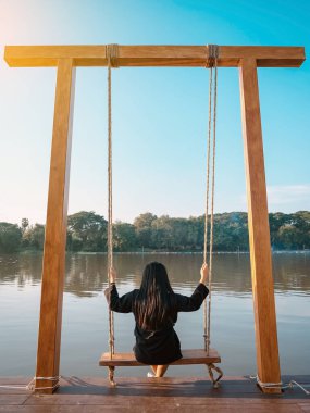 Genç Asyalı kadının arkası rahatça oturur ve nehir kıyısındaki yaz tatilinde ahşap salıncaklarda oynar. Yalnız kız salıncakta tek başına ve doğa manzarasının tadını çıkarıyor güzel büyük bir nehirde akşam vakti.