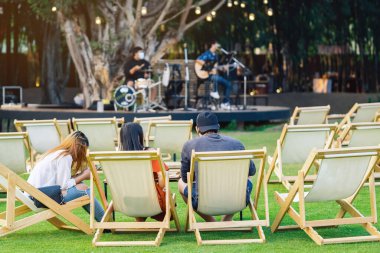 Asyalı genç çiftin arka planı, arka planda müzikal performansın bulanık görüntüsüyle çimenlikte akşam yemeği için masalarla beyaz güverte sandalyelerinde rahatlamak için otururken, gölgeli yeşil çimlerle çevrilidir..