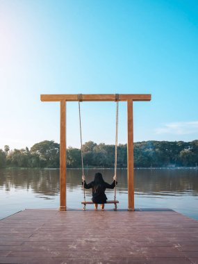 Genç Asyalı kadının arkası rahatça oturur ve nehir kıyısındaki yaz tatilinde ahşap salıncaklarda oynar. Yalnız kız salıncakta tek başına ve doğa manzarasının tadını çıkarıyor güzel büyük bir nehirde akşam vakti.