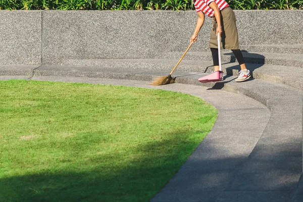 Female worker sweeping yard with broom tool and dustpan in ornamental backyard garden. Woman wear apron with hat sweeping away fallen leaves and sweeps trash. Wooden Broom in hand sweeping up ground.