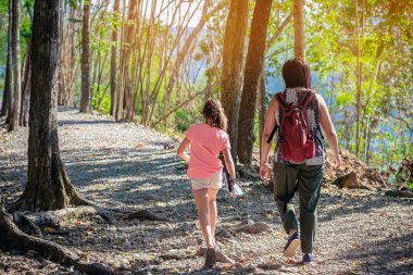 Back view of young girl and her mother walking together in forest nature path walk on trail woods background at Hellfire Pass in Kanchanaburi Thailand. Happy people relaxing on active outdoor activity