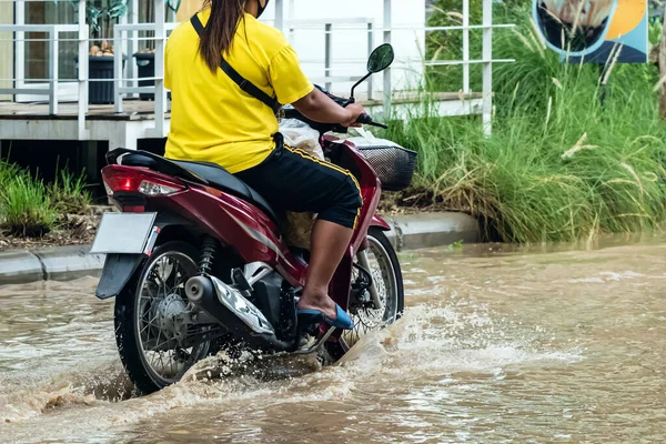 Woman ride motorcycle passing through flooded road. Riding motorbike on flooded road during flood caused by torrential rains. Flooded road with large puddle. Splash by motorcycle through flood water.