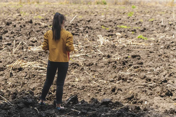 Back view of Asian young woman farmer stand alone to look soil quality for farming in soil field. Female agriculturist think and plan about cultivation in vacant land. Farm and agricultural concept.