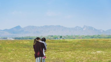 Back view of happiness Asian mother holding daughter with meadow and mountain in background. Lovely Family looking at beautiful sky together. Joy of outdoor living together. Happy family relationship.