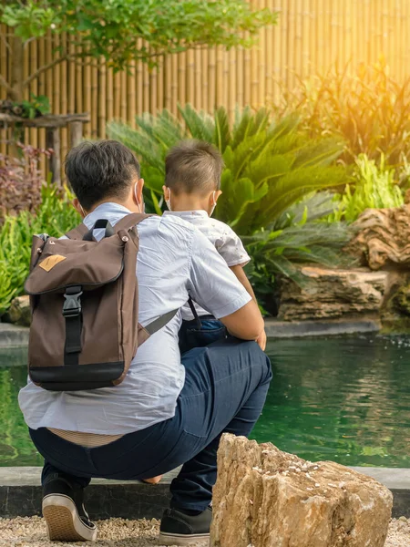Back view of Asian father wear face mask with backpack embraces his son wear mask to watch carp fish in pond decorated in Japanese garden style. Happiness of family living outdoors together concept.