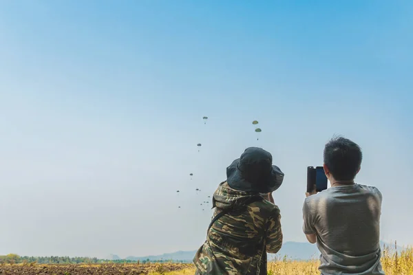 Parents take video clips and photo with smart phone and watch with worry and concern during parachute training from airplane for army cadet with blurred image of parachute and landscape in background.