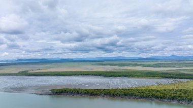 Manzaranın katmanları bir gelgit deresinin üzerinde uçuyor ön planda mangrov ağaçları ve bulutlu bir gökyüzünün altında dağlar var.