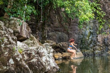 Airlie Beach, Queensland, Australia - January 2022: Girl jumps off rocks into natural swimming hole pool at Cedar Falls