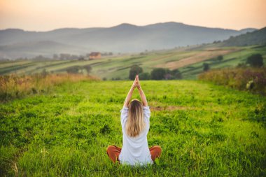 Woman doing yoga on the green grass on the top of the mountain with beautiful view at sunset