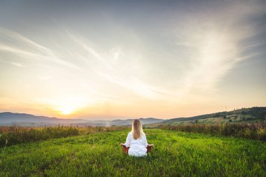 Dağın tepesindeki yeşil çimlerde yoga yapan kadın gün batımında güzel manzaralı.
