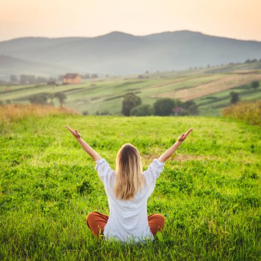 Woman doing yoga on the green grass on the top of the mountain with beautiful view at sunset