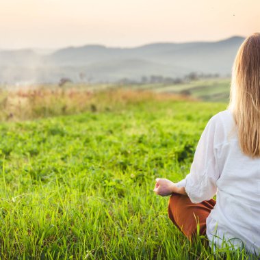 Dağın tepesindeki yeşil çimlerde yoga yapan kadın gün batımında güzel manzaralı.