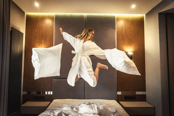 beautiful female droping pilows and jumping with bathrobe on bed in hotel room