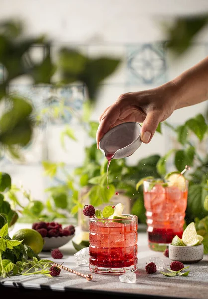 Le donne versano il liquore a mano nel cocktail di lamponi con bacche fresche e lime sul tavolo con succosa verdura.. — Foto Stock