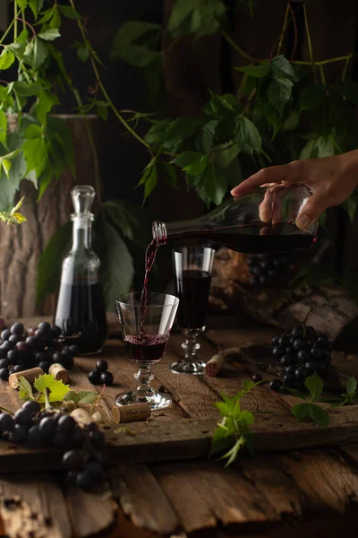 Sürahili kırmızı şarap sahnesi, kristal bardaklar, taze üzümler ve eski ahşap masadaki yeşil dallar. Köy tarzında taze şarap.. — Stok fotoğraf