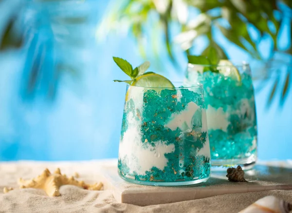 Blaues Schichtendessert mit blauem Curaçao-Gelee, Kokosjoghurt, Limetten- und Minzblättern am Strand auf Marmorplatte mit Seestern-, Muschel- und Palmblättern im Hintergrund. Großaufnahme von geschichtetem süßem Cocktail. — Stockfoto
