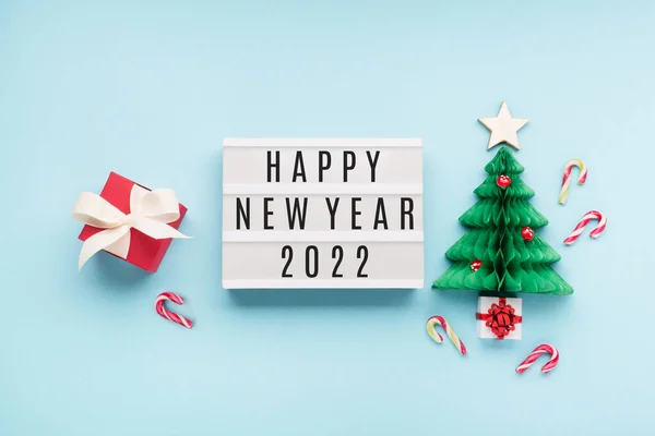 Happy New Year 2022  text on white Lightbox with Christmas paper green tree, mushrooms and red gift box on blue background. Flat lay, top view, copy space. Festive holiday card concept.