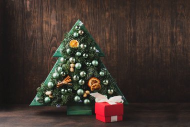 Işıkları, gümüş süslemeleri, portakalları, tarçınları ve koyu ahşap arka planda kırmızı hediye kutusu olan şık ahşap Noel ağacı. Klasik tatil kış festivali kartı konsepti, kopyalama alanı.