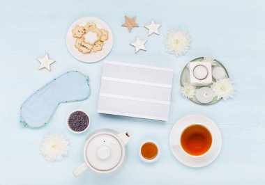 Boş beyaz ışık kutusu, pamuk uyku maskesi, ev yapımı kurabiyeler, bir fincan bitki çayı, kuru lavanta çiçekleri ve mavi arka planda mumlar. Üst manzara, düz yatış. Sağlıklı gece uykusu konsepti.