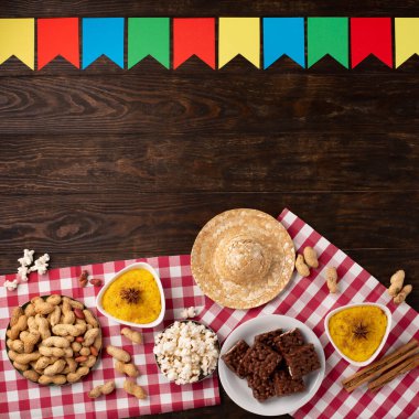 Junina Yaz Festivali Karnavalı konsepti. Brezilya hasır şapkası, patlamış mısır, fıstık ve ahşap arka planda renkli bayraklar, üst manzara. Tebrik Kartı, Davetiye veya Tatil Posteri Tasarımı