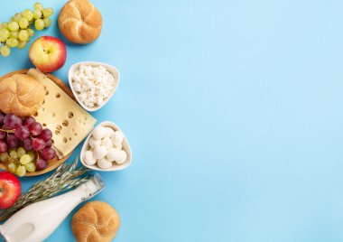 Happy Shavuot festive card, Jewish religious holiday concept. Dairy products, fruits, cheese, bread, milk bottle on blue background. Flat lay, top view, copy space.