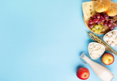 Happy Shavuot festive card, Jewish religious holiday concept. Dairy products, fruits, cheese, bread, milk bottle on blue background. Flat lay, top view, copy space.