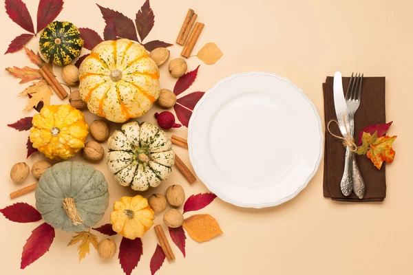 Autumn fall holiday Thanksgiving table place setting. Pumpkins, porcelain white plate, dry maple leaves, spice and apple on beige pastel background. Flat lay, top view, copy space.