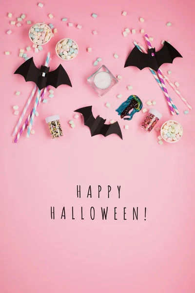 Happy Halloween holiday concept. Halloween decorations, paper black bats, candy on pink background. Halloween party greeting card mockup, copy space. Flat lay, top view, overhead.