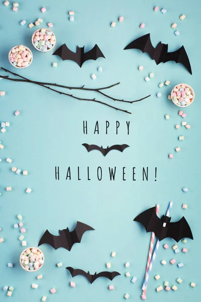 Happy Halloween holiday concept. Halloween decorations, paper black bats, candy on blue background. Halloween party greeting card mockup, copy space. Flat lay, top view, overhead.