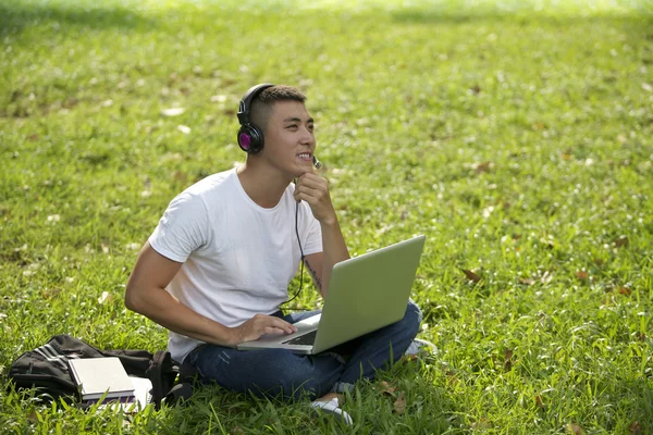 Young handsome Asian student with laptop and headset out door — Stock Photo, Image