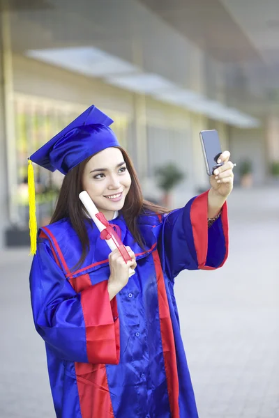 Portrait of a beautiful young asian woman in graduation cap and gown standing outside on campus — Stock Photo, Image