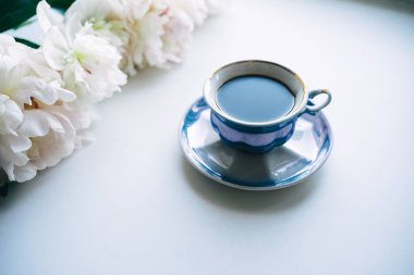 A beautiful postcard. A white coffee cup with a saucer, candles and a vase with a bouquet of white peonies. Beautiful still life. Spring, summer time. The concept of 