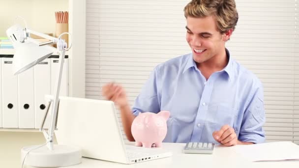 Empresário feliz usando uma calculadora e economizando dinheiro em um banco porquinho - dentro de casa — Vídeo de Stock