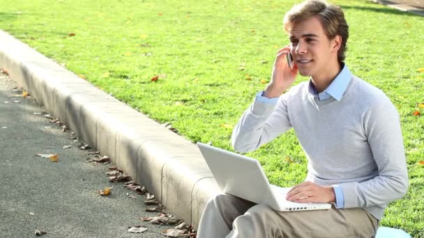 Atractivo joven estudiante es feliz usando el ordenador portátil en el parque - al aire libre — Vídeo de stock